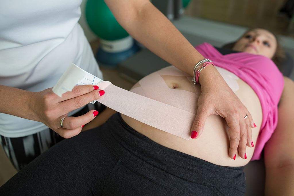 Fizjoterapeuta wykonujący kinesiotaping u kobiety w ciąży. Taping skutecznie wspomaga mięśnie brzucha także w okresie ciąży i połogu.